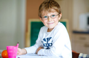 Boy wearing glasses and writing, sitting at a table and smiling at the camera.